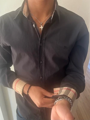 Svart skjorta från Burberry med rutiga detaljer - Stilren svart skjorta från Burberry med klassiska beige-rutiga detaljer på insidan av krage och manschetter. Skjortan har knappar framtill och lång ärm. Perfekt för dig som gillar en clean look med exklusiva detaljer. Inte använd mycket. Den är riktig. 