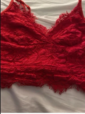 Röd spetsbralette med smala band - Säljer en snygg röd bralette i spets med smala axelband och vacker blommig spetsdetalj. Braletten har en längre modell och dekorativ kant nertill. Perfekt för dig som vill ha något både bekvämt och stilrent under kläderna.