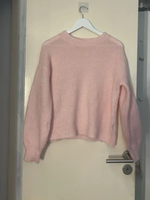 Ljusrosa stickad tröja - Mysig ljusrosa stickad tröja med rund halsringning och långa ärmar. Tröjan har en relaxed passform och ribbade muddar vid ärmslut och nederkant. Perfekt för lager på lager under kyliga dagar.