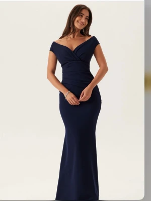 Mörkblå lång klänning - Elegant mörkblå långklänning med off shoulder-skärning och figurnära passform. Klänningen har omlottdetaljer upptill och en lätt utsvängd nederdel som ger en snygg siluett. Perfekt för bal! Slutsåld på hemsidan
