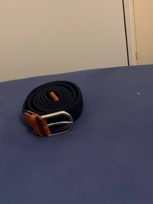 Mörkblått flätat bälte med läderdetaljer - Snyggt mörkblått flätat bälte med bruna läderdetaljer och silverfärgat spänne. Perfekt accessoar för att lyfta din outfit och funkar till både jeans och chinos. Klassisk design som passar till det mesta.