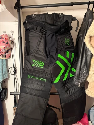 Oxdog X2 innebandybyxor svart/grön - Säljer ett par svarta innebandybyxor från Oxdog, modell X2, med gröna detaljer och loggor. Byxorna har förstärkta partier, justerbar midja med bälte och bred passform för bra rörelsefrihet. Perfekta för målvaktsspel och tuffa matcher.