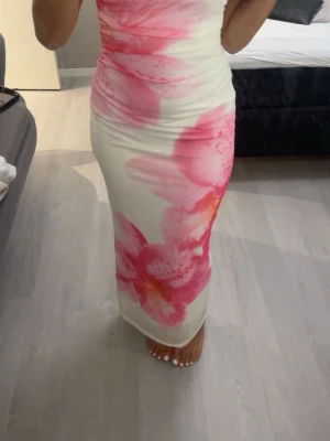 Vit maxiklänning med rosa blommor - Säljer en lång, figurnära maxiklänning i vitt med stora rosa och orange blommönster. Klänningen är axelbandslös och har en mjuk, stretchig passform som framhäver siluetten. Perfekt för sommarens alla tillfällen. Använd en gång.