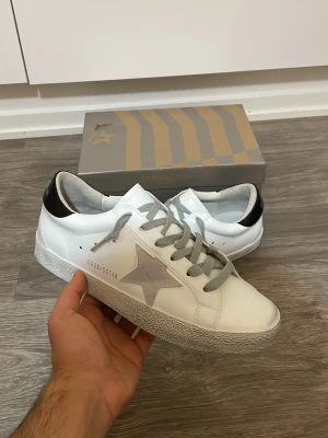 Golden Goose skor - Säljer ett par vita sneakers från Golden Goose med grå stjärna på sidan, grå skosnören och svart detalj på hälen. Skorna har en klassisk låg modell och är tillverkade i läder med slitna detaljer på sulan för en cool look.