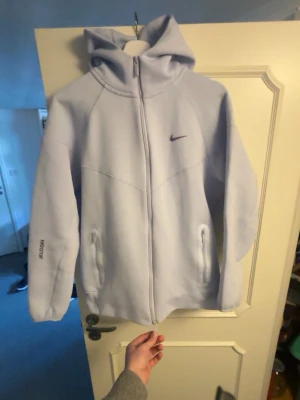 Ljuslila hoodie från Nike x NOCTA - Säljer en ljuslila hoodie från Nike i samarbete med NOCTA. Hoodien har dragkedja framtill, två fickor med dragkedja och en stor huva. Nike-logga på bröstet och NOCTA-tryck på ärmen. Tillverkad i mjukt material som känns riktigt skönt.