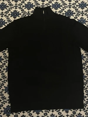 Svart stickad tröja med halv dragkedja - En stilren svart stickad tröja med hög krage och halv dragkedja framtill. Tröjan har långa ärmar och ribbade muddar vid ärmslut och nederkant. Den är i storlek L. Den är använd ett fåtal gånger och är i nästan nyskick. Jag säljer den eftersom den är för stor för mig.  