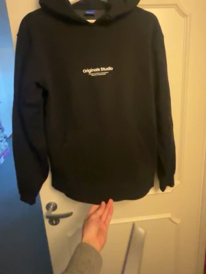 Svart hoodie från Originals Studio - Snygg svart hoodie från Originals Studio med vit text på bröstet. Klassisk modell med huva och känguruficka framtill. Perfekt för en chill och avslappnad stil. Tillverkad i mjukt material som känns skönt mot huden.