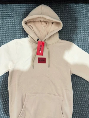 Beige hoodie från Hugo med logga - Snygg beige hoodie från Hugo med röd logga på bröstet och matchande röd tag. Hoodien har huva med snören och en stor magficka. Materialet är mjukt och skönt, perfekt för en avslappnad look. Diskret Hugo-brodyr på ärmen.