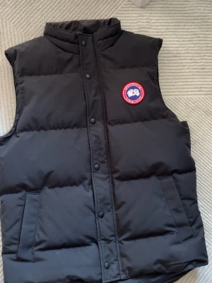 Äkta  Canada Goose väst kvitto finns  -  ÄKTA  svart dunväst från Canada Goose med klassisk röd och vit logga på bröstet. Västen har hög krage, tryckknappar framtill och två fickor. Perfekt för lager-på-lager och håller dig varm under kyliga dagar. KVITTO FINNS priset är ej hugget i sten 