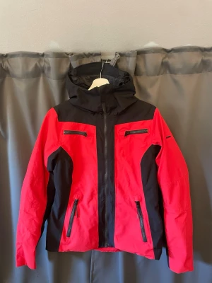 Röd och svart skid/vindjacka med huva - Snygg skid/vindjacka i rött och svart med dragkedjor framtill och två bröstfickor. Jackan har en huva och svarta detaljer längs sidorna och ärmarna. Perfekt för blåsiga dagar och outdoor-aktiviteter. Materialet känns slitstarkt och vattenavvisande. Även lite figursydd