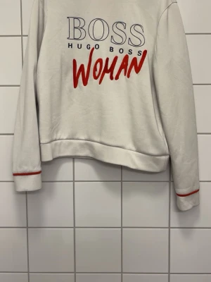 Vit sweatshirt från Hugo Boss Woman - Vit sweatshirt från Hugo Boss Woman med tryck i blått och rött framtill. Tröjan har rund hals, långa ärmar och röda detaljer vid ärmsluten. Tillverkad i mjukt material som känns skönt mot huden. Perfekt för en avslappnad och trendig look.