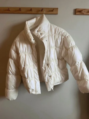 Vit kort pufferjacka med hög krage - Säljer en snygg vit pufferjacka i kort modell med hög krage och dragkedja samt tryckknappar framtill. Jackan har en puffig siluett och breda kanaler som ger en modern look. Perfekt för kalla vinterdagar och riktigt trendig just nu.
