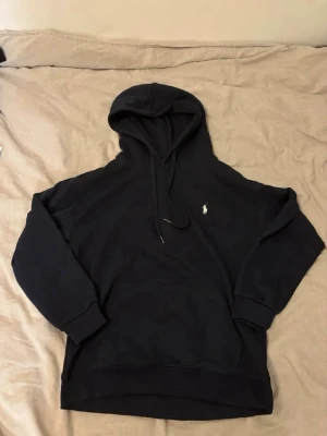 Blå hoodie från Polo Ralph Lauren - Säljer en blå hoodie från Polo Ralph Lauren med klassisk luva och dragkedja. Tröjan har det ikoniska vita Polo-märket broderat på bröstet och ribbade muddar vid ärmar och nederkant. Perfekt för en clean och stilren look.
