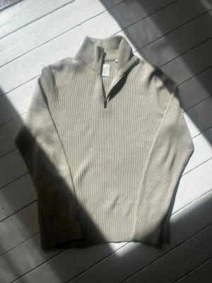 Beige ribbad halvzip tröja - Snygg beige stickad tröja med ribbad struktur och halvzip i halsen. Tröjan har lång ärm och hög krage, perfekt för lager på lager. Enkel och stilren design som passar till det mesta.