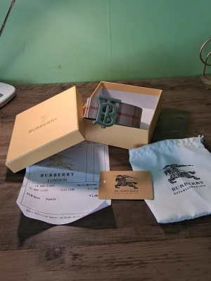 Burberry rutig bälte med silverdetalj - Snyggt Burberry-bälte med klassiskt rutigt mönster i beige, rött och grått. Bältet har ett stort silvrigt B-spänne framtill och levereras med originalbox och dustbag. Perfekt accessoar för att lyfta din outfit. Denna är lite använd men det är inget seriöst med den.