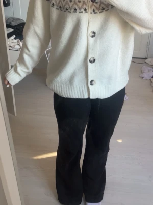 Vit kofta med mönster och knappar - Mysig vit stickad kofta med stora knappar framtill och ett klassiskt nordiskt mönster i beige, grått och svart över bröstet. Koftan har ribbade muddar och en avslappnad passform, perfekt för lager på lager under kyliga dagar.