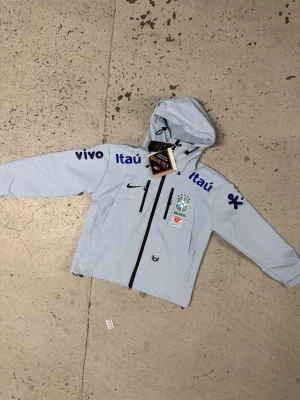 Nike Brasil vindjacka med huva - Ljusblå vindjacka från Nike med Brasilien fotbollslandslagets logga på bröstet och flera sponsorloggor i blått. Jackan har huva, hel dragkedja framtill, två bröstfickor med dragkedja och två sidofickor. Tillverkad i GORE-TEX-material för extra väderskydd.