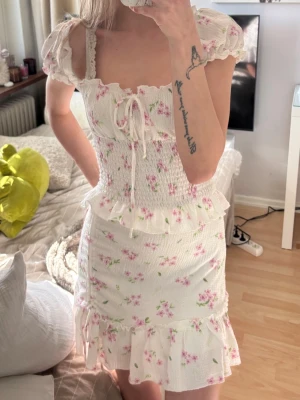 Vit blommig kortklänning med volang - Supersöt vit kortklänning med rosa blommönster, puffärmar och volangdetaljer. Klänningen har smockad midja, knytning fram och volanger vid ärmslut och kjolkant. Perfekt för sommarens alla tillfällen.