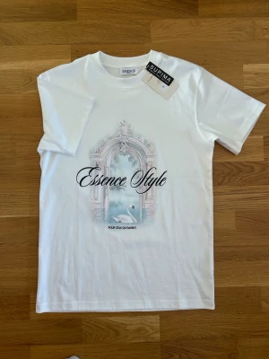 Vit Essence Style t-shirt med tryck - Snygg vit t-shirt från Essence med trycket 'Essence Style' och ett motiv av en svan vid ett klassiskt valv. Den är helt ny och perfekt nu till sommaren!
