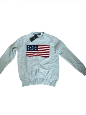 Ljusblå stickad tröja Polo Ralph Lauren - Ljusblå stickad tröja från Polo Ralph Lauren med amerikansk flagga i rött, vitt och blått på bröstet. Rund halsringning och ribbade muddar vid ärmslut och nederkant. RL-broderi under flaggan. Perfekt för dig som gillar klassisk preppy stil.
