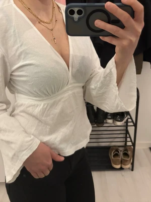 Vit topp från Gina Tricot - Säljer en vit omlottopp från Gina Tricot i storlek M. Toppen har v-ringning, vida ärmar och är gjord i ett lätt, strukturerat bomullsliknande material. Perfekt att styla med jeans eller kjol för en trendig look.