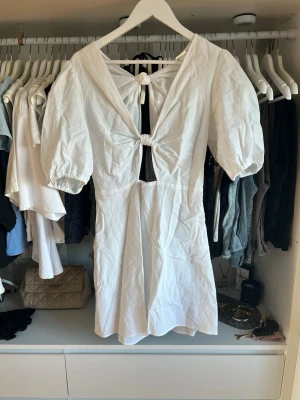 Vit kortklänning med puffärm och knyt - Trendig vit kortklänning med puffärmar och två knyt framtill och baktill som ger en öppen rygg-effekt. Klänningen har en v-ringad urringning och är tillverkad i ett lätt bomullstyg, perfekt för varma dagar. Snygg och luftig modell som känns modern och fräsch.