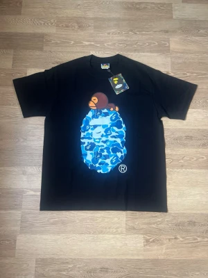 Svart BAPE t-shirt med blått camomönster - Säljer en svart t-shirt från BAPE med ikoniskt blått camomönster och apa-tryck på bröstet. Klassisk passform och rund hals. Perfekt för dig som gillar streetwear och vill sticka ut med en unik design.