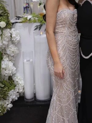 Glittrig långklänning i champagne - Säljer en stunning glittrig långklänning i champagnefärg med pärlbroderier och paljettdetaljer. Klänningen är axelbandslös och har en elegant, figurnära passform med släp. Perfekt för bal eller festliga tillfällen. Materialet känns lyxigt och skimrar i ljuset. Är en 36 men insydd till 34/32. Släpet är avtagbart.