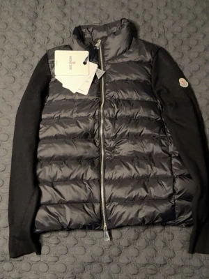 Svart cardiganjacka från Moncler - Snygg svart pufferjacka från Moncler med quiltad front och rygg i glansigt material samt stickade svarta ärmar. Jackan har hög krage, dragkedja framtill och Moncler-logga på ärmen. Perfekt för dig som vill ha en stilren och trendig jacka. STORLEK PASSAR S. HELT NY OANVÄND.