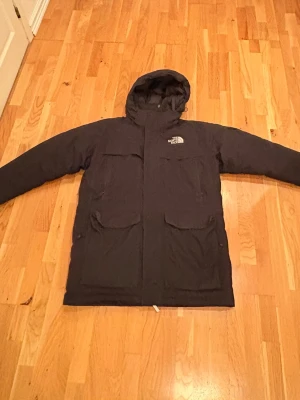 North Face jacka  - Svart vinterjacka från The North Face med huva och stora fickor framtill. Klassisk logga på bröstet och ryggen. Jackan är lång och har en rak passform, perfekt för kalla dagar. Materialet är slitstarkt och håller värmen bra.