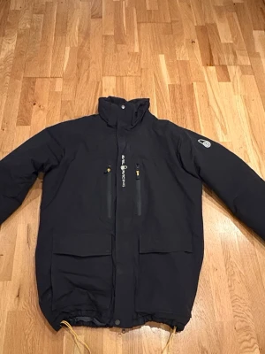 Svart vindjacka från Sail racing - Säljer en svart vindjacka från Sail Racing med hög krage, två bröstfickor med dragkedja och två stora framfickor. Jackan har justerbar dragsko nedtill och logga på ärmen och ryggen. Materialet är slitstarkt och jackan har en clean look utan synlig huva.