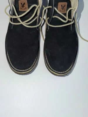 Svarta mockakängor från Lyle & Scott - Snygga svarta kängor i mocka från Lyle & Scott med beige snörning och kontrastsömmar. Klassisk design med rundad tå och diskret logga på plösen. Perfekta för dig som vill ha stilrena boots med en modern touch.