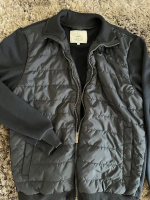 Svart quiltad cardiganjacka från Bläck - Snygg svart cardiganjacka från Bläck med quiltad front och mjuka stickade ärmar. Jackan har dragkedja framtill, ribbade muddar och två fickor. Perfekt för lager på lager och en clean look.