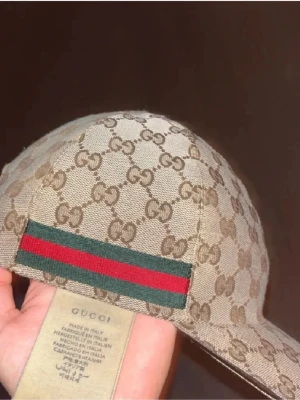 Beige gg keps - Säljer en beige keps  med det klassiska GG Supreme-mönstret i brunt över hela kepsen. På sidan finns den ikoniska gröna och röda randen. Kepsen är tillverkad i canvasmaterial och har böjd skärm. Perfekt för dig som vill ha en lyxig och trendig accessoar.