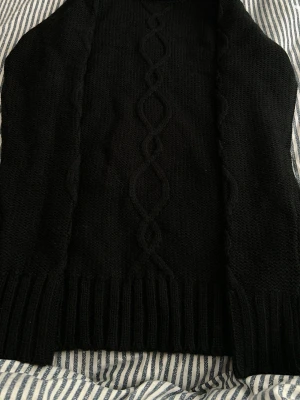 Svart stickad tröja med flätmönster - En svart stickad tröja med snyggt flätmönster framtill och ribbade muddar vid nederkant. Tröjan har en klassisk rak passform och är perfekt för kyligare dagar. Materialet ser ut att vara mjukt och värmande, perfekt för lager på lager.