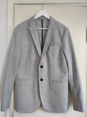 Ljusgrå blazer från Zara - Stilren ljusgrå blazer från Zara med två knappar framtill och klassisk krage. Kavajen har två framfickor och en bröstficka, samt mörkblått innerfoder. Perfekt för en trendig och clean look.