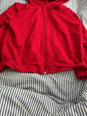 Röd hoodie med dragkedja - Röd hoodie med dragkedja framtill och två stora fickor. Klassisk modell med huva och ribbade muddar vid ärmslut och nederkant. Tillverkad i mjukt bomullstyg som känns skönt mot huden. Perfekt för en avslappnad och stilren look.