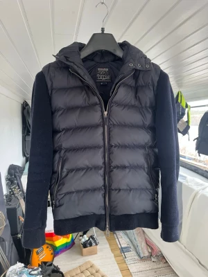 Woolrich cardiganjacka  - Navy cardiganjacka från Woolrich med vadderad kropp och stickade ärmar. Jackan har dragkedja framtill, två fickor med dragkedja och en stor huva. Sitter mycket bra på kroppen och är en bra riktigt fet cardigan nu för våren och sommaren. Finns en liten defekt på baksidan av jackan längst ned som nu e lappad (se sista bilden), påverkar ingenting och syns heller inte i helhet när den är på! Annars super skick, hör av er vid frågor eller funderingar! 
