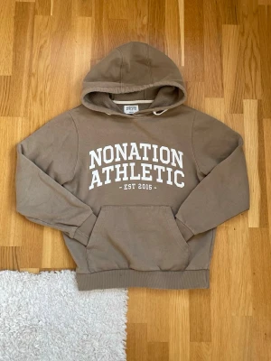 Beige hoodie från Nonation Athletic - Snygg beige hoodie från Nonation Athletic med stor vit text på bröstet och känguruficka framtill. Klassisk huva med dragsko och ribbade muddar vid ärmslut och nederkant. Perfekt för en avslappnad och sportig stil.