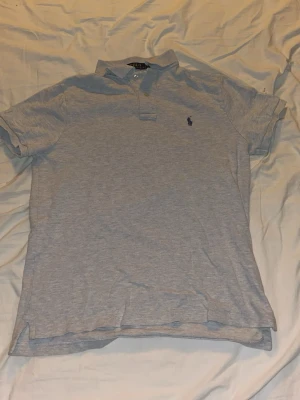 Ljusgrå pikétröja från Polo Ralph Lauren - Klassisk ljusgrå pikétröja från Polo Ralph Lauren med blå logga broderad på bröstet. Tröjan har korta ärmar, krage och knappar vid halsen. Modellen är custom slim fit och passar perfekt till en avslappnad men stilren look.