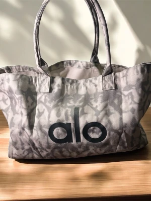 Grå shoppingväska från Alo - Snygg shoppingväska i grått med batikinspirerat mönster och stort Alo-tryck på framsidan. Väskan har dubbla handtag och är tillverkad i slitstarkt tygmaterial. Perfekt för att bära med sig allt man behöver under dagen.