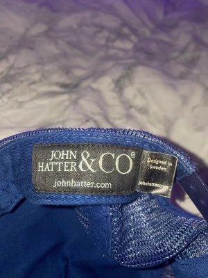 Blå keps John Hatter & Co THE KING - Snygg blå keps från John Hatter & Co med mesh på sidorna och baksidan. Framtill sitter en gul skylt med texten 'THE KING' i svart. Justerbar passform och klassisk böjd skärm. Perfekt för dig som vill sticka ut med en cool accessoar.