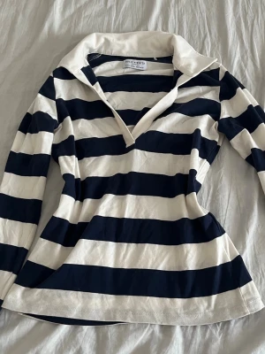 Randig långärmad topp från Holly and whyte - Snygg långärmad topp från Hollister med breda marinblå och vita ränder. Toppen har krage och v-ringad öppning framtill. Tillverkad i mjuk bomull som känns skön mot huden. Perfekt för en avslappnad och trendig look.