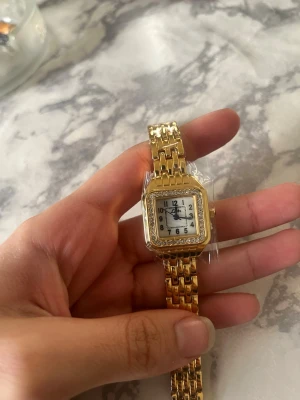 Guldfärgad fyrkantig klocka - Stilren guldfärgad klocka med fyrkantig urtavla och vit bakgrund. Klockan har en länkad metallrem och tydliga svarta siffror på urtavlan. Perfekt accessoar för dig som gillar klassisk och elegant stil. Den kommer med en grej där man kan ändra storleken på klockan ifall den inte passar dig och den är helt ny från ur och Penn som jag köpte för ett tag sen som jag it använder eftersom jag har andra klockor som jag har dagligen och har ej tid för den här och färgen går ej bort och helt oanvänd. 