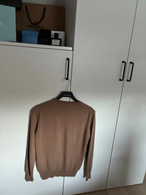 Brun tröja från J.Lindeberg - Snygg brun tröja från J.Lindeberg med diskret logga broderad på bröstet. Tröjan har rund halsringning, långa ärmar och ribbade muddar. Perfekt för dig som gillar stilrena och klassiska plagg.