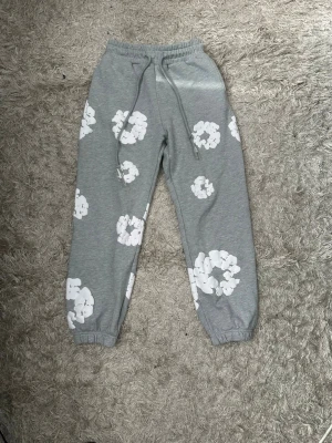 Grå denim tears mjukisbyxor  - Säljer ett par grå denim tears mjukisbyxor med vita blom tryck. Byxorna har resår i midjan och benslut samt dragsko i midjan för skön passform. Perfekta för chill dagar hemma eller när du vill ha en avslappnad stil. Passar perfket med den matchande hoodien
