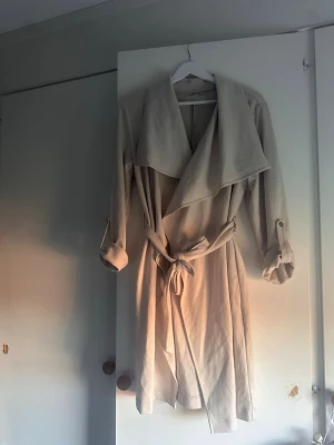 Beige trenchcoat med knytskärp - Stilren beige trenchcoat med bred krage och knytskärp i midjan. Jackan har långa ärmar med dekorativa slejfar vid ärmsluten och ett mjukt, följsamt tyg som ger en avslappnad look. 