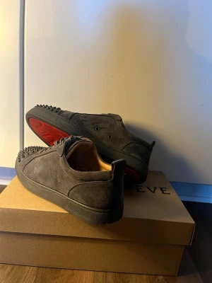 Gråa sneakers med nitar från Louboutin - Säljer ett par gråa sneakers i mocka från Christian Louboutin. Skorna har coola nitar på tån, matchande grå skosnören och en låg siluett. Insidan är beige och sulan är svart. Perfekta för dig som vill sticka ut med en edgy look. Kan även gå ner i pris vid snabbaffär