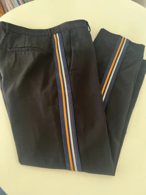 Svarta byxa med ränder Samsøe Samsøe - Snygga svarta byxa  från Samsøe Samsøe med blå, gula och vita ränder längs sidorna. Klassisk passform med bälteshällor och bakficka. Perfekta för dig som vill ha en unik twist på dina byxor.