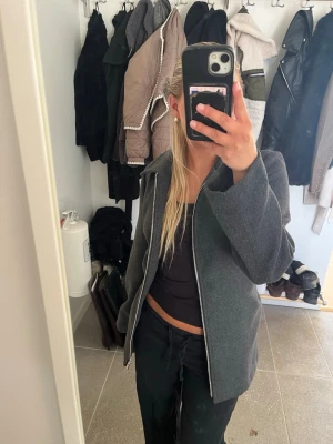 Kappa - Stilren mörkgrå kappa med rak passform, dragkedja framtill och tillhörande skärp i midjan. 